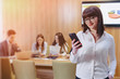 © pressahotkey - Stylish office worker woman in glasses with phone in hands against background of working colleagues