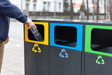 Hand Of Man Throwing Plactic Bottle Into Trash Bin For Plastic Waste. Modern Colorful Refuse Bins For Separate Trash.