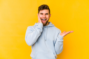 Wall Mural - Young caucasian man isolated on yellow background holds copy space on a palm, keep hand over cheek. Amazed and delighted.