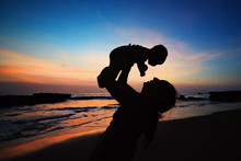 Happy Family Black Silhouette On Sunset Sky Background. Young Mother, Baby Son Have Fun Together, Walk By Sea Beach. Mom Toss And Catch Child. Travel Lifestyle, Parents With Kids On Summer Holidays.