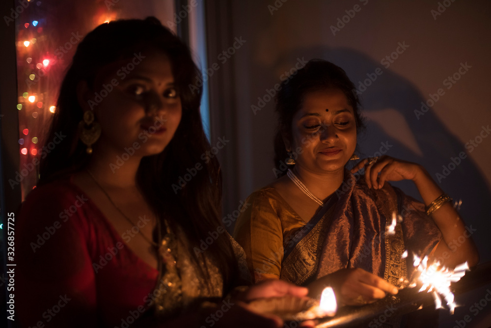 Two young and beautiful Indian Bengali women in Indian traditional dress are celebrating Diwali with diya/lamp and fire crackers on a balcony in darkness. Indian lifestyle and Diwali celebration - obrazy, fototapety, plakaty 