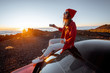 © rh2010 - Young woman dressed in red enjoying sunset landscape above the clouds, sitting on the car roof while travel in the mountains. Carefree lifestyle and travel concept