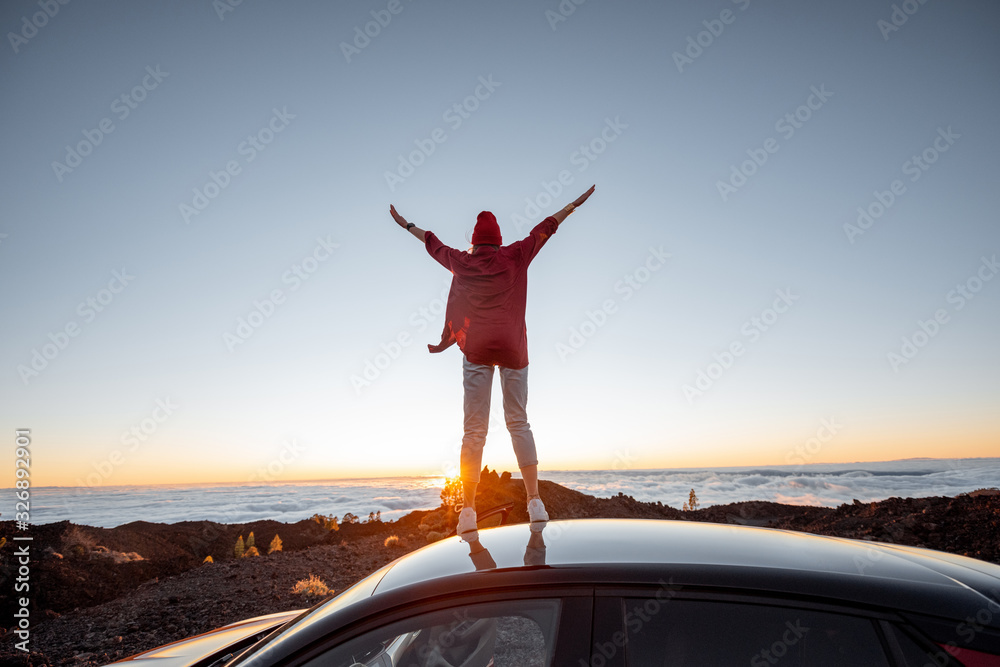 Woman enjoying rocky landscapes above the clouds on a sunset, standing on the car highly in the mountains. Carefree lifestyle and travel concept - obrazy, fototapety, plakaty 