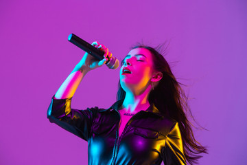 Wall Mural - Caucasian female singer portrait isolated on purple studio background in neon light. Beautiful female model in black wear with microphone. Concept of human emotions, facial expression, ad, music, art.