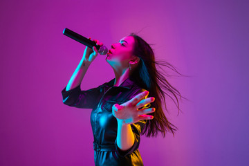 Wall Mural - Caucasian female singer portrait isolated on purple studio background in neon light. Beautiful female model in black wear with microphone. Concept of human emotions, facial expression, ad, music, art.