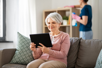 Wall Mural - technology, old age and people concept - happy senior woman with tablet pc computer and housekeeper cleaning at home