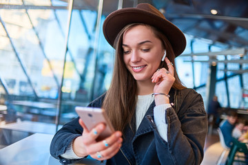 Wall Mural - Stylish fashionable happy hipster woman in hat listen music online using mobile phone and wireless white headphones in a cafe. Modern people with digital lifestyle