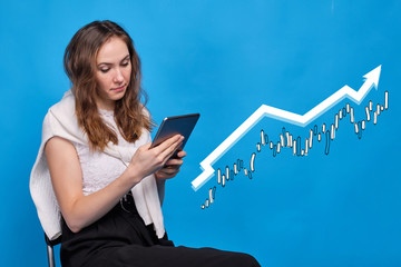 A girl in casual clothes on a blue background plays successfully on the stock exchange through a tablet. Abstract chart and growth arrow.