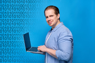 A man in casual clothes on a blue background looks at the camera and programs on a laptop. Binary code abstract.