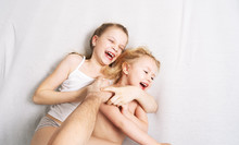 Family Time: Dad Tickles His Daughters. The Girls Are Lying On The Bed And Laughing Merrily.