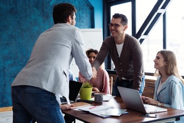 Wall Mural - Welcome to our team! Young modern men in smart casual wear shaking hands while working in the creative office.