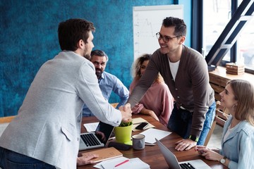 Wall Mural - Welcome to our team! Young modern men in smart casual wear shaking hands while working in the creative office.