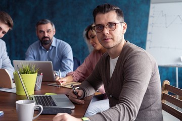 Happy to be a part of great team. Cheerful young handsome man looking at camera with smile while sitting at the office table on business meeting with his coworkers.