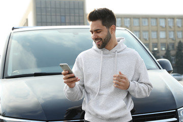 Handsome young man with smartphone near modern car outdoors