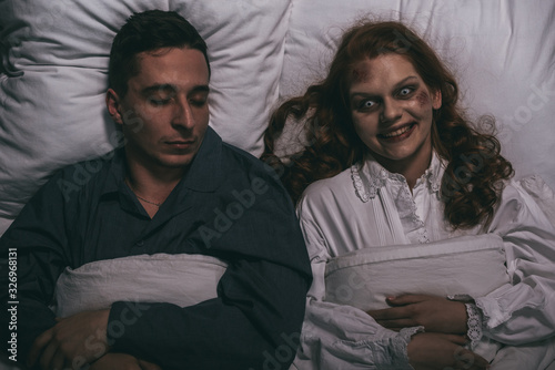 Cuadros en Lienzo top view of creepy smiling female demon lying in bed with sleeping man