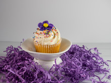 Vanilla Cupcake With White Frosting And A Purple Flower And Sprinkles On A White Dish Surrounded By Purple Paper Shred.  Festive, Pretty And Delicious!