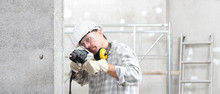 Man Using An Electric Pneumatic Drill Making A Hole In Wall, Professional Construction Worker With Safety Hard Hat, Hearing Protection Headphones, Gloves And Protective Glasses. 