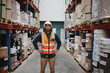 © StratfordProductions - Manager standing in warehouse between shelf filled with goods wearing a white helmet and orange vest for protection with hands on waist
