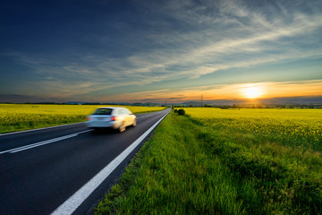 Wall Mural - motion blurred white car driving on the empty asphalt road between the yellow flowering rapeseed fie