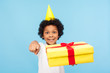 © khosrork - Funny excited joyful little boy with party cone on head pointing to camera and holding gift box, child choosing you, congratulating on birthday and giving awesome present. studio shot blue background