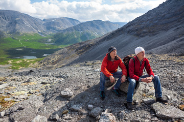 Caucasian father with his adult son sitting and resting on stone when climbing to the height of the mountain