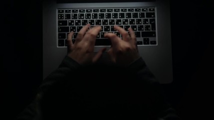 Overhead hacker in the hood working with laptop and mobile phone typing text in the darkroom. Russian hacker hacking the server in the dark. A hacker girl sitting in the dark with her laptop