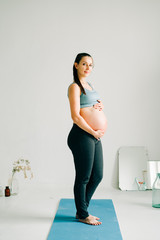 Wall Mural - a young pregnant girl in sportswear is standing on a yoga Mat, smiling and holding her belly in the Studio