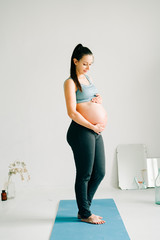 Wall Mural - a young pregnant girl in sportswear is standing on a yoga Mat, smiling and holding her belly in the Studio