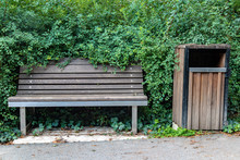An Empty Wooden Bench With A Waste Bin Beside Way With The Green Bush Around.
