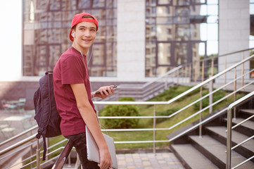 Smiling teenager holds cell phone and laptop, in the city.  Young man stands with a smartphone and computer, in the street.  Lifestyle concept of a happy teenage boy, outdoors.