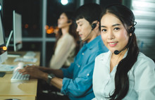 Asian Confidence Operator Woman Agent With Headsets Working In A Call Center At Night Environment With Her Colleague Team As Customer Service.Successful Business People In Headsets Are Using Gadgets .