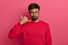 Serious Looking Unshaven European Man Makes Call Me Back Gesture, Keeps Always In Touch, Wears Transparent Spectacles And Red Sweater, Asks For Telephone Number, Isolated On Pink Background.