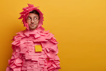Indoor Shot Of Male Advertiser Or Manager Covered With Sticky Adhesive Notes, Looks Aside As Notices Something Interesting, Poses Against Yellow Background, Free Space For Your Advertising Content