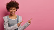 Young delighted curly African American woman has cheerful expression, points aside with fore fingers, shows blank space for your advertising content, wears grey jacket, poses over pink background
