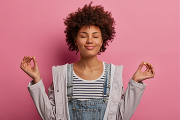 Portrait of relieved determined dark skinned woman meditates with closed eyes, practices breathing exercise or yoga to calm down, stands in lotus pose, poses over pink background, makes zen gesture