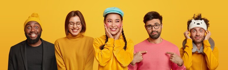 Cheerful two women smile toothily, one man points at himself, other listens audio track in headphones, wears sleep mask, dark skinned guy in yellow hat and black suit, pose indoor over yellow wall