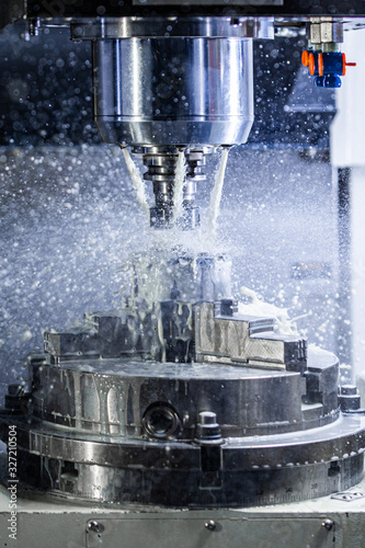 Vertical photo of industrial wet milling process in 5-axis cnc machine with coolant flow under pressure and freezed splashes