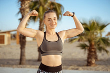 Strong Sporty Woman Showing Her Biceps Muscles On The Beach With Palm Tree On Background.