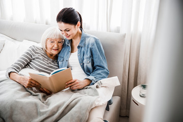 Wall Mural - Joyful old woman reading book with adult granddaughter