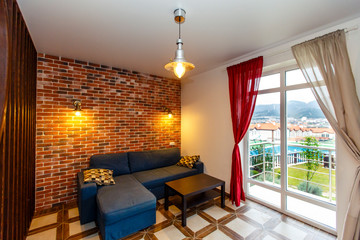 A blue sofa with a coffee table in the back of the cottage. Behind the sofa is a decorative wall of old red brick with two lamps. Open window with red and white curtains-mountain view