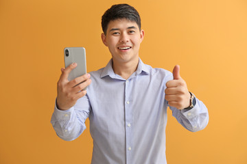 Poster - Young Asian businessman with mobile phone showing thumb-up on color background