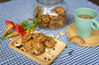 chocolate chip and nuts cookies decorated on a wooden plate with a cup of coffee, a cookie jar and flowers on a blue and white checked table cloth