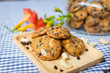 closeup of chocolate chip and nuts cookies decorated on a wooden plate with a cookie jar and flowers on a blue and white checked table cloth