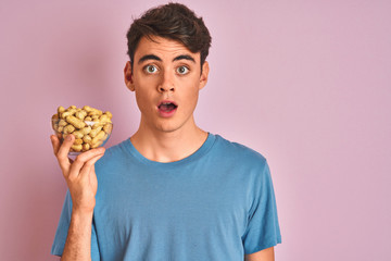 Sticker - Teenager boy holding a bunch of peanunts over isolated pink background scared in shock with a surprise face, afraid and excited with fear expression