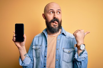 Sticker - Handsome bald man with beard holding smartphone showing screen over yellow background pointing and showing with thumb up to the side with happy face smiling