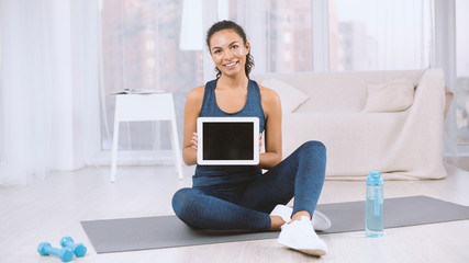 Happy Latin woman showing tablet computer with empty screen on yoga mat at home, mockup for design