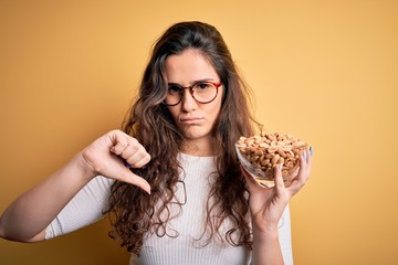 Poster - Beautiful woman with curly hair holding bowl with healthy peanuts over yellow background with angry face, negative sign showing dislike with thumbs down, rejection concept