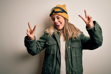 Canvas Print - Young brunette skier woman wearing snow clothes and ski goggles over white background smiling with tongue out showing fingers of both hands doing victory sign. Number two.