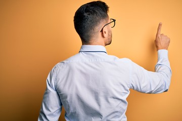 Young handsome businessman wearing tie and glasses standing over yellow background Posing backwards pointing ahead with finger hand