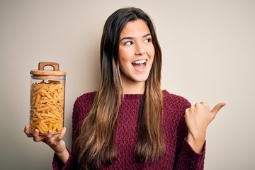 Canvas Print - Young beautiful girl holding bottle of dry Italian pasta macaroni over white background pointing and showing with thumb up to the side with happy face smiling